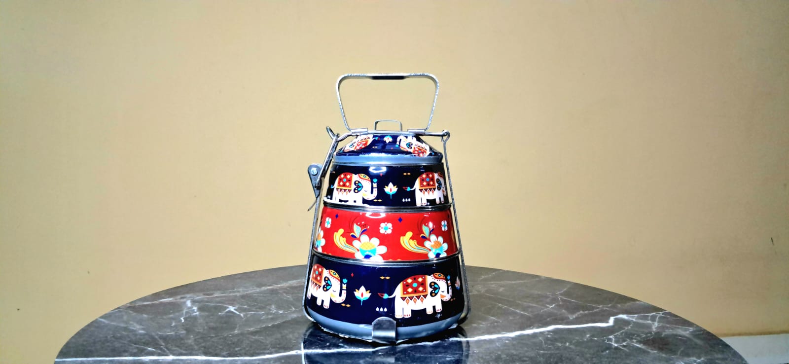 Colorful tiered hand painted Tiffin with animal designs placed on a marble surface against a beige wall.